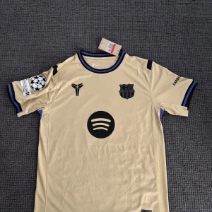 Beige FC Barcelona Rashford Nike tröja - Snygg beige FC Barcelona fotbollströja från Nike med svarta detaljer, Rashford 14 på ryggen och Spotify-logga på bröstet. Kortärmad modell i Dri-FIT material, perfekt för träning eller match. Champions League-märke på ärmen och klubbmärke på bröstet.