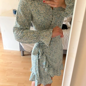 Blommig omlottklänning Rut&Circle XS - Supersöt kort omlottklänning från Rut&Circle i blått med små vita och gula blommor. Klänningen har långa ärmar, volangkant nertill och knyt i midjan. Perfekt för vår och sommar när du vill känna dig extra fin.