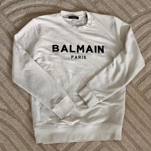 Vit sweatshirt från Balmain Paris - Vit sweatshirt från Balmain Paris med svart logotyptryck på bröstet. Klassisk rund halsringning, långa ärmar och ribbade muddar. Tillverkad i mjuk bomull för en skön och avslappnad look.