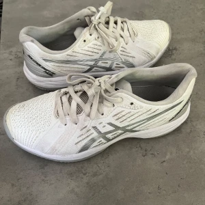 Vita Asics sneakers strl 37,5 - Säljer ett par vita Asics sneakers med silverdetaljer och snörning. Ovandelen är i mesh och syntet för bra andningsförmåga och komfort. Sulan har ett mönstrat grepp och skorna passar perfekt för träning eller sportiga outfits.