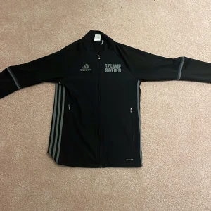 Svart Adidas Camp Sweden jacka XS - Svart Adidas jacka i sportig stil med Camp Sweden-tryck på bröstet och flera sponsorer på ryggen. Jackan har hel dragkedja, två stora rymbara fickor med dragkedja och grå ränder längs sidorna. Tillverkad i lätt och ventilerande polyester, perfekt för dig som gillar sportig look.