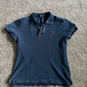 Marinblå pikétröja från Ralph Lauren - Klassisk marinblå pikétröja från Ralph Lauren med röd broderad logga på bröstet. Modellen är 'The Skinny Polo' med korta ärmar, krage och knappar framtill. Perfekt för en stilren och sportig look.