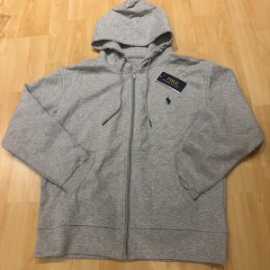 Grå hoodie från Polo Ralph Lauren - Snygg grå hoodie från Polo Ralph Lauren med dragkedja, huva och snörning. Klassisk logga broderad på bröstet och två fickor framtill. Perfekt för en chill och stilren look.