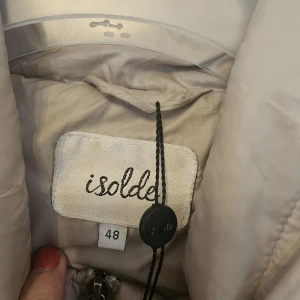 Beige quiltad pufferjacka Isolde - Snygg beige pufferjacka från Isolde med quiltad design och dragkedja framtill. Jackan har höga krage, två fickor med dragkedja och långärmade ärmar. Perfekt för dig som gillar stilren och enkel look med lite volym.