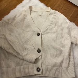 Beige stickad kofta från H&M - Mysig beige stickad kofta från H&M med stora, snygga knappar framtill. Koftan har v-ringning, ribbade muddar och en relaxed passform. Perfekt att slänga på sig över en t-shirt eller linne när det blir lite kyligare.