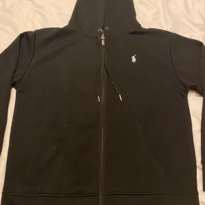 Svart hoodie från Polo Ralph Lauren - Svart hoodie från Polo Ralph Lauren i bomull med dragkedja framtill, snörning i huvan och klassisk vit logga på bröstet. Slim fit-modell med känguruficka och stilren look. Perfekt för dig som gillar enkel och snygg streetstyle.