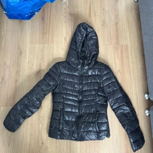 Svart pufferjacka från ONLY XS - Svart pufferjacka från ONLY i storlek XS. Jackan har huva med dragsko, horisontella sömmar och dragkedja framtill. Perfekt för kyliga dagar och enkel att matcha med olika outfits.