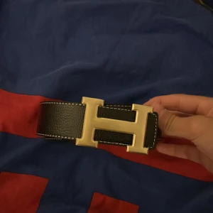 Svart Her mes bälte med guldspänne - Säljer ett svart bälte från Her mes med ett stort, ikoniskt H-spänne i guld. Bältet är gjort i äkta skinn med vit kontrastsöm längs kanterna. Perfekt accessoar för att lyfta vilken outfit som helst.