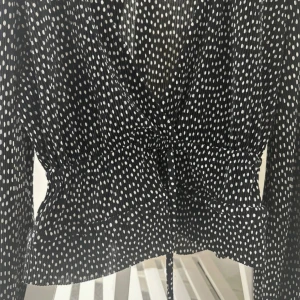 Prickig omlottblus från Zara - Svart omlottblus från Zara med vita prickar, v-ringning och långa ärmar. Blusen har knyt i midjan och lätt volang nedtill. Tunt och luftigt material som ger en snygg siluett. Perfekt för dig som gillar trendiga mönster och en feminin look.