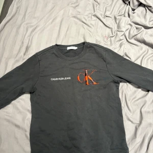 Svart långärmad tröja Calvin Klein - Svart långärmad tröja från Calvin Klein Jeans med stort broderat CK-logga i rött och svart på bröstet. Tröjan har rund halsringning och är tillverkad i mjuk bomull. Perfekt för dig som gillar stilrena plagg med en cool twist.