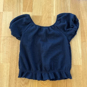 Mörkblå blus med puffärm - Supersöt mörkblå blus med puffiga korta ärmar och smockad struktur. Blusen har en rundad halsringning och volangkant nertill som ger en romantisk vibe. Perfekt att styla med jeans eller kjol för en trendig look.