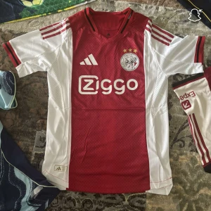 Ajax fotbollströja Adidas röd/vit - Ajax fotbollströja från Adidas med klassisk röd mittpanel och vita sidor och ärmar. Tröjan har korta ärmar, tre röda ränder på axlarna och klubbmärke samt Ziggo-logga på bröstet. Tillverkad i lätt och ventilerande material, perfekt för match eller träning.