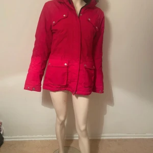 Röd parkajacka med huva - Riktigt snygg röd parkajacka med huva och dragkedja framtill. Jackan har fyra fickor med lock och tryckknappar, samt justerbara ärmslut. Perfekt för dig som vill ha en färgstark jacka med praktiska detaljer och skön passform. Jackan är helt ny oanvänd med har inte prislapp 