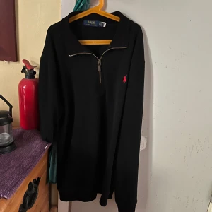 Svart half zip tröja Polo Ralph Lauren - Svart långärmad tröja från Polo Ralph Lauren med röd broderad logga på bröstet. Tröjan har en snygg half zip-dragkedja vid halsen och är gjord i 100% bomull. Perfekt för dig som gillar klassisk och stilren streetwear.