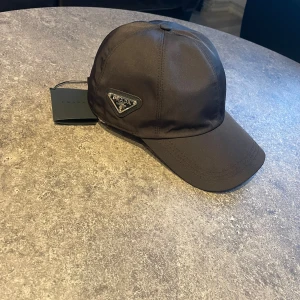 Svart keps från Prada i nylon - Snygg svart keps från Prada med klassisk böjd skärm och ikonisk metalltriangel med logga på sidan. Tillverkad i slät nylon som ger en stilren och modern look. Perfekt accessoar för dig som gillar exklusiva märken och minimalistisk design.