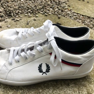 Vita Fred Perry sneakers med röd/blå detalj - Snygga vita sneakers från Fred Perry i textil med klassisk laurel-logga på sidan. Skorna har vita snören och en röd/blå rand längs sidan. 