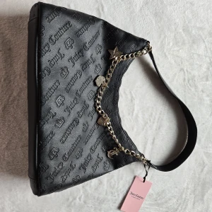 Svart axelväska Juicy Couture med kedja - Snygg svart axelväska från Juicy Couture i präglat skinnimitation med logotyp över hela väskan. Väskan har en guldfärgad kedja med flera coola berlocker som stjärna och hjärta. Perfekt accessoar för att lyfta din outfit.