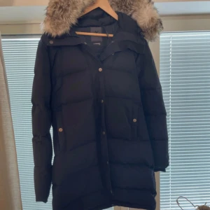 Svart dunjacka med fuskpäls från Everest - Lång svart dunjacka från Everest med huva och snygg fuskpäls runt kragen. Jackan har dragkedja och knappar framtill, två fickor med lock och en klassisk pufferlook. Perfekt för kalla vinterdagar och riktigt mysig att bära.