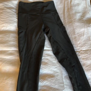 Svarta högmidjade leggings från Maya delorez  - Svarta leggings från Maya delorez med hög midja och diskret mönstrade partier längs benen. Tillverkade i stretchigt syntetmaterial, perfekta för ridning. Snygg passform och praktiska fickor på sidorna. De har ett litet hål ner på smalbenet men igen man ser när de är på, men påverkar ju såklart priset!