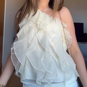 Vit volangig oneshoulder blus - Säljer en superfin vit blus med volanger och oneshoulder-design. Toppen har en luftig och romantisk look med flera lager av tunna volanger som ger extra rörelse. Perfekt för dig som gillar att sticka ut med en unik och feminin stil.