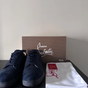Mörkblå sneakers från Christian Louboutin - Snygga mörkblå sneakers från Christian Louboutin i mocka med klassisk röd sula och rund tå. Skorna har snörning och en stilren design som sticker ut med den ikoniska röda undersidan. Kommer med originalkartong och dustbag.