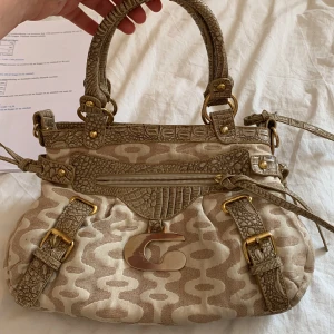 Beige handväska från Guess med mönster - Snygg beige handväska  med ikoniskt G-spänne i metall, mönstrad canvas och detaljer i krokopräglat skinn. Väskan har guldfärgade metalldetaljer, dragkedja och justerbara remmar. Perfekt accessoar för att lyfta din outfit. finns lite fläckar 