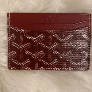 Bordeaux Goy4rd plånbok med mönster - Säljer en bordeauxfärgad plånbok från Goy4rd med deras ikoniska vita och bruna grafiska mönster. Plånboken är i skinn och har flera kortfack. Kommer med originalask, dustbag och kvitto. Perfekt för dig som vill ha något lyxigt och stilrent i fickan.