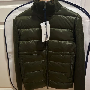 Moncler Cardigan - Snygg olivgrön dunväst från Moncler med stickade ärmar och rygg. Västen har hög krage, hel dragkedja framtill och två fickor med dragkedja. Framsidan är quiltad och glansig medan ärmar och rygg är ribbstickade för en cool kontrast.