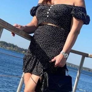 Svart prickig offshoulder klänning - Supersöt svart kortklänning med vita prickar och offshoulder-modell. Klänningen har puffiga korta ärmar, knappar framtill och volang nertill. Perfekt för varma dagar och ger en trendig vibe.