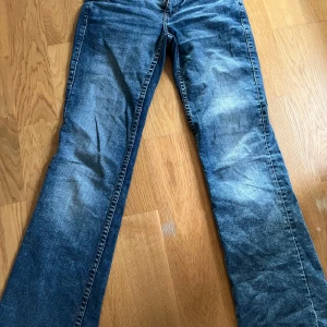 Blå bootcut jeans med fickdetaljer - Snygga blå jeans med bootcut-modell och klassisk femficksdesign. Jeansen har en lätt tvättad look och raka ben som blir vidare nedtill. Bakfickorna har lock och knapp, vilket ger en extra detalj. Perfekta för dig som gillar en avslappnad men trendig stil.