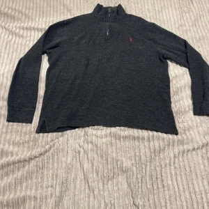 Mörkgrå half zip tröja från Ralph Lauren - Mörkgrå långärmad tröja från Ralph Lauren med half zip och hög krage. Tröjan har en liten röd broderad logga på bröstet och är tillverkad i mjuk bomull. Perfekt för lager-på-lager och chill stil. Storlek L – passar även M