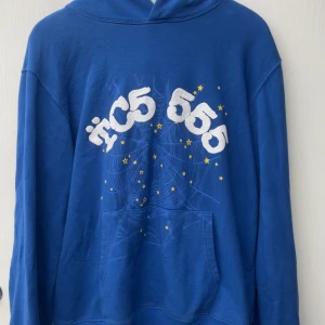 Blå hoodie från sp5der - Blå hoodie med vit och gul tryckt design framtill, där siffrorna 555 och stjärnor syns över ett spindelnätsmönster. Huva och känguruficka framtill. Ärm med gul detalj och små stjärnor. Perfekt för dig som gillar streetwear och coola prints.
