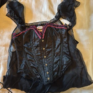 Svart korsett från Buttricks med snörning  - Säljer en svart korsett från Buttricks med snygga detaljer i lila och rosa längs överkanten. Korsetten har knäppning fram och snörning bak för perfekt passform. Materialet är glansigt och känns lyxigt, med volangkant upptill som ger en extra cool look.