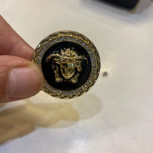  brosch med Medusa-huvud - Säljer en rund brosch  med Medusa-huvud i guld på svart bakgrund. Runt kanten sitter gnistrande stenar som ger extra bling. Broschen är i metall med tydlig logga på baksidan och säkerhetsnål för enkel fastsättning.