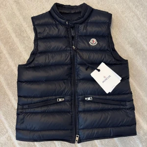 Mörkblå dunväst från Moncler - Snygg mörkblå dunväst från Moncler med quiltad design och ståkrage. Västen har två dragkedjeförsedda fickor framtill, Moncler-logga på bröstet och en smidig dragkedja i mitten. Perfekt för lager-på-lager och stilrena outfits. OBS dragkedjan är av som ni kan se på bild 3!