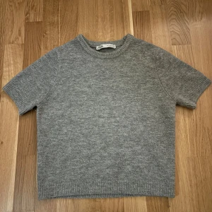 Grå stickad tröja från Zara - En enkel och stilren grå stickad tröja från Zara i storlek S. Tröjan har korta ärmar, rund hals och ribbade kanter vid ärmslut och nederkant. Perfekt för lager-på-lager och chill vibes under höst och vinter. Något nopprig men inget man riktigt tänker på vid användning❤️