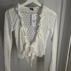 Vit blus med volang från Gina  - Supersöt vit omlott blus från gina i storlek XS. Blusen har ett snyggt geometriskt mönster, långa ärmar och volangkant längs ringningen. Knytband framtill och lätt transparent material   