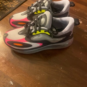 Nike Air Max 720 sneakers färgglada - Nike Air Max 720 sneakers med futuristisk design, mesh-överdel i grått och svart, detaljer i neonrosa, gul och orange. Snygga svarta snören och genomskinlig plattformssula med maxad dämpning. Perfekt för dig som gillar att sticka ut med färg och stil.