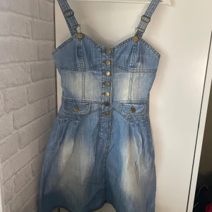 Jeansklänning med knappar från H&M - Snygg vintage jeansklänning från H&M i ljusblå denim med justerbara axelband och dekorativa mässingsknappar framtill. Klänningen har fakefickor på sidorna och knappar mitt på som går att knäppa upp. Perfekt för dig som gillar denim.