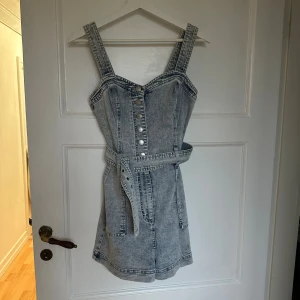 Jeansklänning med bälte från FB Sister - Snygg ljusblå jeansklänning från FB Sister i kort modell. Klänningen har breda axelband, knappar framtill, markerad midja med tillhörande bälte och stora fickor både fram och bak. Perfekt för en cool och avslappnad look.