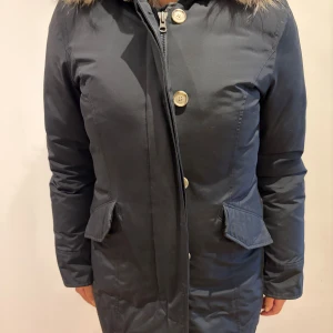 Svart dunjacka från Woolrich med päls - Snygg svart dunjacka från Woolrich med huva och fluffig pälsdetalj. Jackan har långa ärmar, dragkedja och knappar framtill samt två fickor med lock. Perfekt för kalla dagar och har en stilren, lång passform.