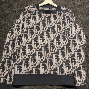 Mönstrad stickad tröja från Dior - Säljer en stickad tröja från Dior i marinblått och beige med ett coolt all-over logomönster. Tröjan är i 100% ull och har ribbade muddar vid hals, ärmslut och nederkant. Perfekt för dig som vill sticka ut med en lyxig statement-piece.