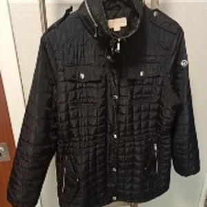 Svart quiltad jacka Michael Kors - Svart quiltad jacka från Michael Kors med hög krage, tryckknappar och dragkedja framtill. Jackan har fickor med dragkedja och bröstfickor med lock. Snyggt rutmönster och emblem på ärmen. Perfekt för kyliga dagar.