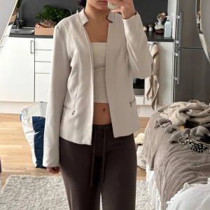 Beige kavaj med dragkedjor - Snygg beige kavaj med stilren design och coola silverfärgade dragkedjor vid fickorna. Jackan har lång ärm, rak passform och en modern krage utan slag. Perfekt för dig som gillar minimalistisk och clean stil.. Står storlek 40 men passar s