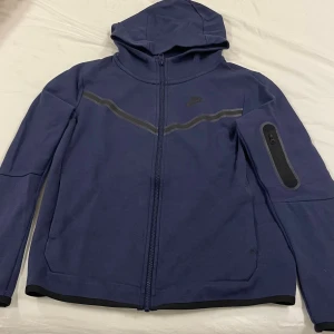 Blå Nike teck  - Nike teck i marinblått med huva och hel dragkedja. Jackan har en snygg svart detalj över bröstet och en praktisk ficka med dragkedja på ena ärmen. Perfekt för dig som gillar sportig stil och vill ha något bekvämt och funktionellt. 158-170 PRISET KAN DISKUTERAS 