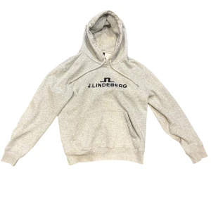 Grå hoodie från J.Lindeberg - Snygg ljusgrå hoodie från J.Lindeberg med svart logotyp på bröstet. Tröjan har huva med snörning, magficka och ribbade muddar. Tveka inte att ställa en fråga!