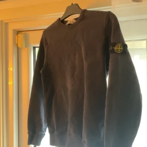 Svart sweatshirt från Stone Island - Svart sweatshirt från Stone Island med klassisk rund hals och ikonisk logga på vänster ärm. Tröjan har ribbade muddar och är tillverkad i mjuk bomull, perfekt för en clean och stilren look.