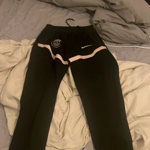 Svarta PSG träningsbyxor från Nike - Svarta träningsbyxor från Nike med Paris Saint-Germain-logga och rosa swoosh på framsidan. Byxorna har rosa detaljer längs benen och snörning i midjan. Tillverkade i polyester, perfekt för dig som gillar sportig stil och fotboll.