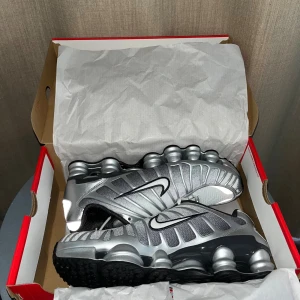 Nike Shox silver sneakers strl 44 - Nike Shox sneakers i silver och svart med futuristisk design och synliga fjädrande Shox-element i sulan. Ovandel i mesh och syntet, svart innersula och vita snören. Snyggt kontrasterande Swoosh på sidan och reflekterande detaljer bak. Perfekt för dig som gillar tech-vibe.