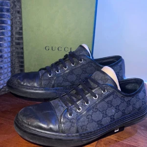 Svarta sneakers från Gucci - Säljer ett par svarta Gucci sneakers med klassiskt GG-monogram i tyg och detaljer i skinn. Skorna har rund tå, platt sula och snörning framtill. Snygg och stilren design som sticker ut med diskret mönster längs sidorna.